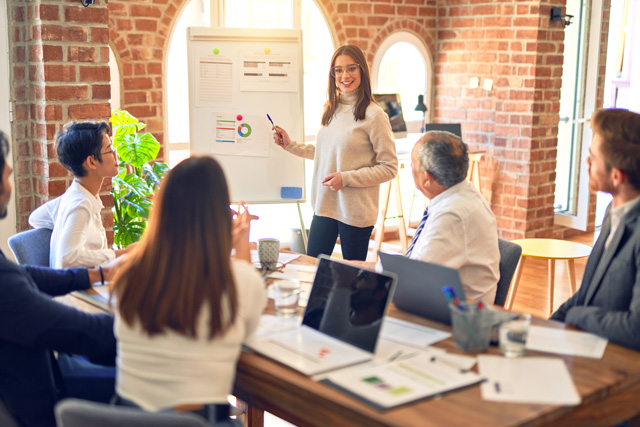 Eine Dozierende steht vor einer Gruppe von Menschen an einer Schautafel. Das Bild steht symbolisch für die Arbeit des Teams hinter der digitalen Baugenehmigung.